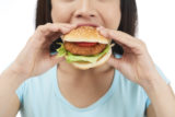 Close-up shot of young woman wearing blue T-shirt eating appetizing hamburger while standing against white background